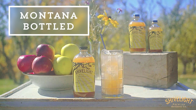 May include: A glass of iced tea with a straw, a bottle of Shivelight Bitteroot Apple & Cinnamon Syrup, and a bowl of red and green apples on a white wooden table. The text "MONTANA BOTTLED" is in a white rectangle on a green background.