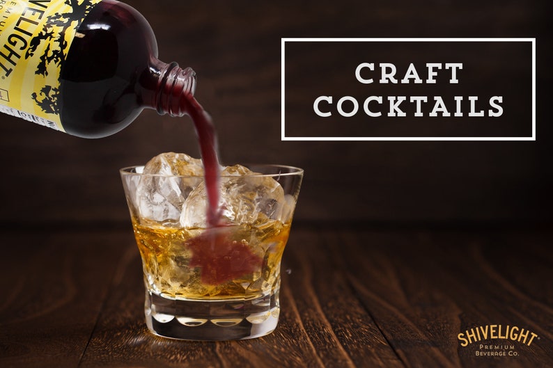 May include: A glass of amber liquid with ice cubes is being poured with a dark red liquid from a bottle labeled "Shivelight Premium Beverage Co.". The text "CRAFT COCKTAILS" is displayed in a white rectangle on a dark brown background.