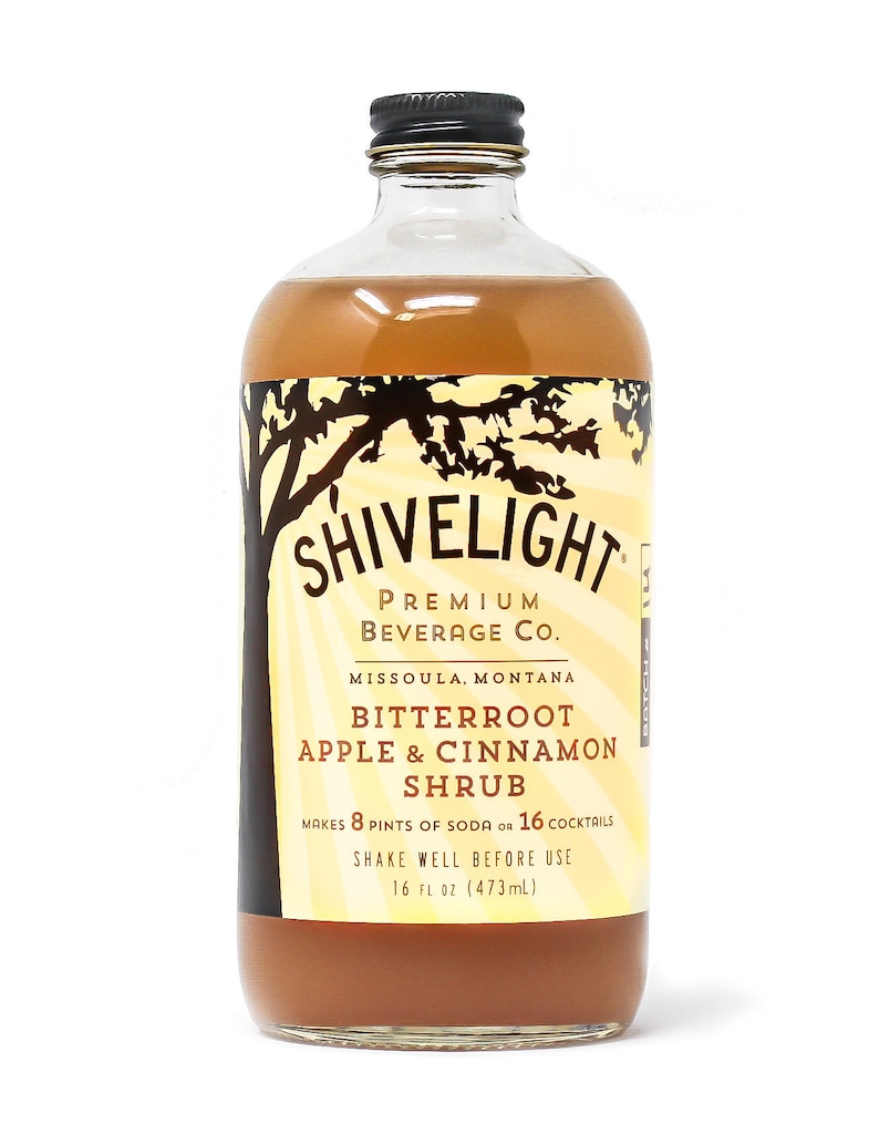 May include: A glass bottle of Shivelight Bitterroot Apple & Cinnamon Shrub, a premium beverage company based in Missoula, Montana. The bottle is filled with a light brown liquid and has a black label with white text. The label states that the shrub makes 8 pints of soda or 16 cocktails. It also says to shake well before use. The bottle contains 16 fluid ounces (473 ml).