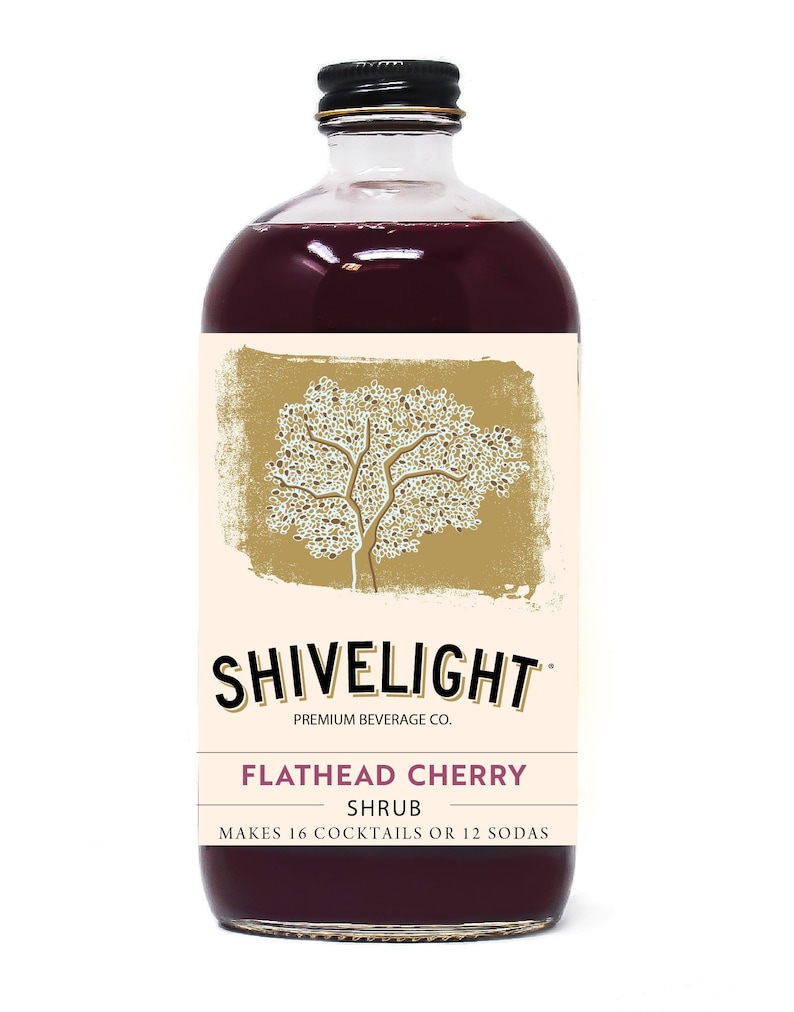 May include: A glass bottle of Shivelight Flathead Cherry Shrub, a premium beverage. The bottle is filled with a dark red liquid and has a black cap. The label features a stylized tree design and the text "Shivelight Premium Beverage Co. Flathead Cherry Shrub Makes 16 Cocktails or 12 Sodas".