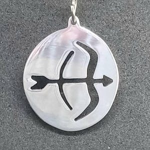May include: A silver pendant necklace featuring a circular pendant with a cut-out design of an archer's bow and arrow. The pendant hangs from a delicate silver chain. The background is a dark gray surface.