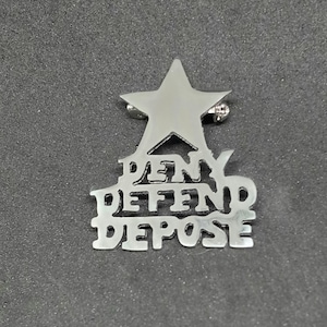 May include: A silver-toned metal pin with a star at the top and the words "Deny Defend Depose" stacked below.
