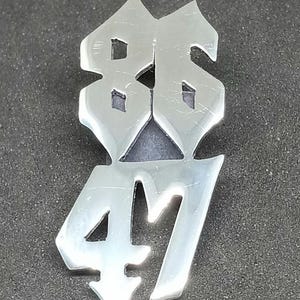 Puede incluir: Un colgante de metal plateado con los números "86" sobre "47" en una fuente gótica estilizada. Los números están conectados y tienen una superficie reflectante, sobre un fondo gris oscuro.