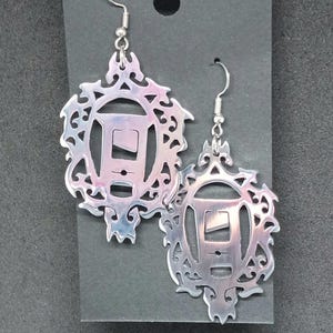 May include: A pair of silver earrings with a detailed, ornate design. Each earring features a circular cutout with a smaller rectangular cutout inside. The earrings have a shiny, metallic finish.