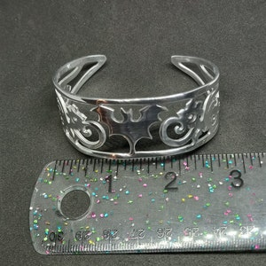 Puede incluir: Una pulsera de plata con un dise&ntilde;o recortado de murci&eacute;lagos y espirales. La pulsera tiene un di&aacute;metro de aproximadamente 5 cm.