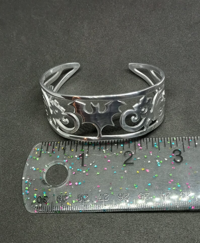 Puede incluir: Una pulsera de plata con un dise&ntilde;o recortado de murci&eacute;lagos y espirales. La pulsera tiene un di&aacute;metro de aproximadamente 5 cm.