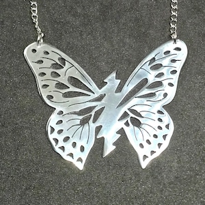 Puede incluir: Un collar con colgante de mariposa plateada. El diseño de la mariposa presenta intrincados detalles recortados en las alas y el cuerpo, suspendido de una delicada cadena. El collar se muestra sobre un fondo oscuro.