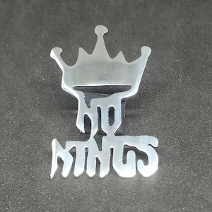 May include: A silver-coloured metal pin in the shape of a crown with the word "KINGS" written below it in stylised lettering. The pin is set against a dark grey background.