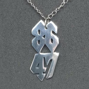 Puede incluir: Un collar con colgante plateado que muestra los números "86" sobre "47" en una fuente estilizada. El colgante cuelga de una delicada cadena. El collar se exhibe sobre un fondo gris oscuro.