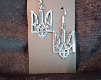 Puede incluir: Un par de pendientes de plata con un diseño de tridente detallado. Los pendientes cuelgan de una tarjeta marrón.