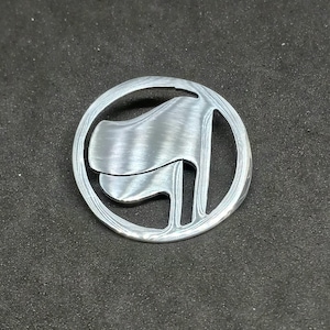 Puede incluir: Un pin de metal plateado con una imagen estilizada de una bandera en un círculo.