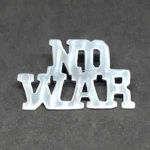 Puede incluir: Un pin metálico plateado con las palabras "NO WAR" recortadas en una fuente audaz y sans-serif. El pin está sobre un fondo gris oscuro, creando un fuerte contraste. El diseño es simple pero impactante.