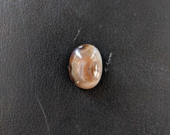 May include: A brown and white agate cabochon with a swirling pattern. The stone is oval shaped and has a smooth, polished surface.