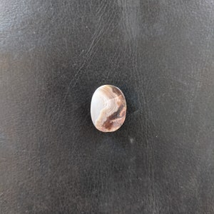 May include: A close-up of a light brown agate cabochon with white and brown banding. The cabochon is oval shaped and has a smooth, polished surface.