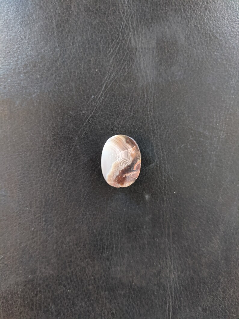 May include: A close-up of a light brown agate cabochon with white and brown banding. The cabochon is oval shaped and has a smooth, polished surface.