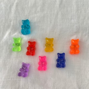 Op de afbeelding: Een verzameling van acht kleurrijke gummy bear snoepjes in verschillende tinten blauw, groen, rood, geel, oranje, paars en roze. De gummy bears zijn gerangschikt op een wit, getextureerd oppervlak.
