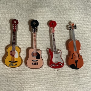 Puede incluir: Cuatro instrumentos musicales en miniatura, incluyendo una guitarra acústica amarilla con la palabra "Mark" en ella, una guitarra acústica rosa con la palabra "NAME" en ella, una guitarra eléctrica roja con la palabra "Mark" en ella y un violín marrón.