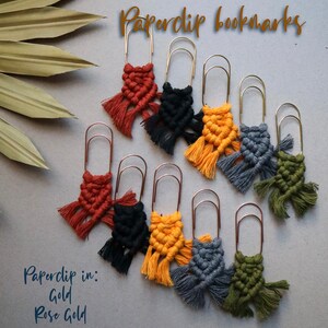 May include: A set of nine colorful macrame bookmarks with tassels attached to gold and rose gold paperclips. The bookmarks are in various colors, including red, black, yellow, gray, and green. The text "Paperclip bookmarks" and "Paperclip in: Gold Rose Gold" is visible in the image.