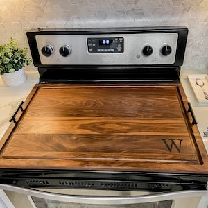May include: A walnut wood stovetop cover with a black handle on each side. The cover has a personalized letter "W" engraved on the surface. The cover is sitting on top of a black and silver stove.