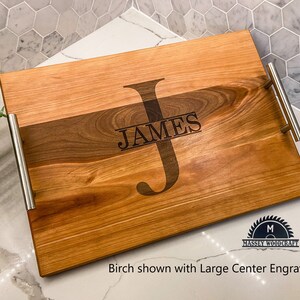 May include: A wooden serving tray with two silver handles. The tray is made of birch wood and has a large center engraving that says "JAMES" with a large "J" in the center. The tray is shown on a white countertop.