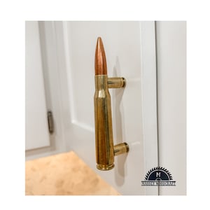 May include: A brass bullet casing door handle with a copper tip. The handle is mounted on a white cabinet door. The handle is a unique and stylish way to add a touch of rustic charm to any home.