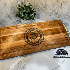May include: A wooden serving tray with brass handles. The tray has a wood-burned design of the United States Marine Corps emblem. The tray is on a white marble surface. The text "Massey Woodcraft" is visible in the lower right corner of the image.