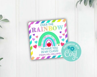 May include: A colorful square card with the text "OVER THE RAINBOW" and "TO HAVE YOU IN MY CLASS!" with a rainbow and apple graphic. The card is decorated with hearts and pencils. The text "FROM: MS. HOLLAND" is also included.