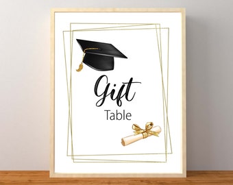 May include: A white sign with gold frames and the text "Gift Table" in black. A black graduation cap with a gold tassel and a gold ribbon tied around a scroll are on the sign.