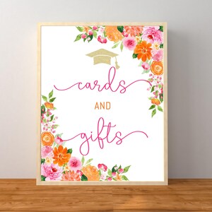 May include: A framed sign with the words "cards and gifts" in pink script, accented by a gold graduation cap and watercolor floral arrangements in shades of pink, orange, and green.