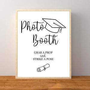 May include: A framed sign with the words "Photo Booth" in elegant script, topped with a graduation cap illustration. Below, it reads "Grab a prop and strike a pose," with a scroll illustration. The sign is set against a white background.