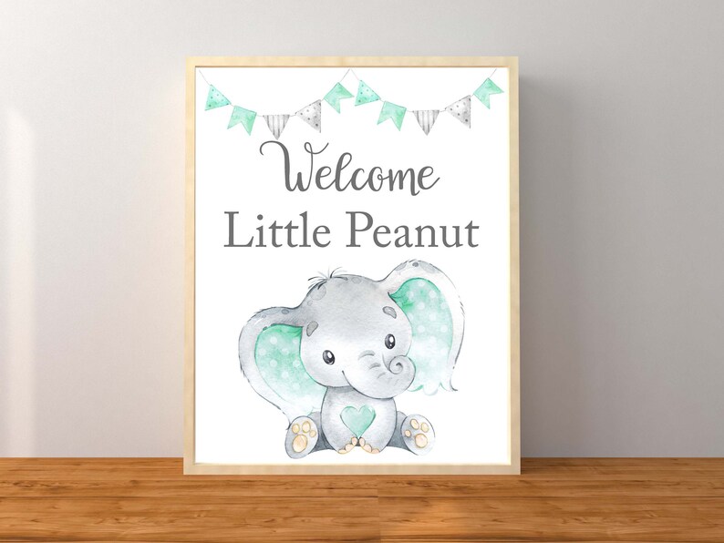 May include: A framed print with a cute cartoon elephant with green polka dots on its ears. The print says "Welcome Little Peanut" and has a string of flags above the elephant.