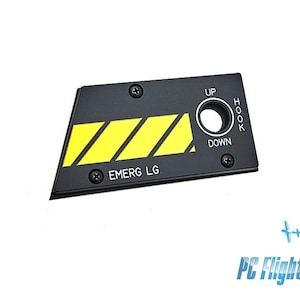 Może przedstawiać: Czarny panel samolotu z żółtymi paskami ostrzegawczymi i tekstem "EMERG LG". Panel ma okrągły pokrętło z tekstem "UP" i "DOWN" oraz prostokątny otwór oznaczony jako "HOOK".