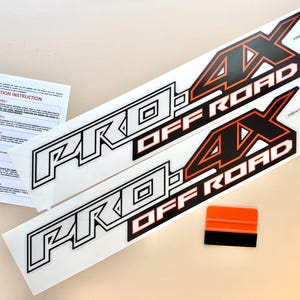 May include: Two white and black "PRO 4X OFF ROAD" decals with orange accents and an orange squeegee. Installation instructions are included. The decals are designed for off-road vehicles.