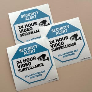 May include: Three blue and white security alert signs with the text "24 Hour Video Surveillance" and a black and white image of a security camera. The text "All Activities Are Monitored" is also included on the signs.