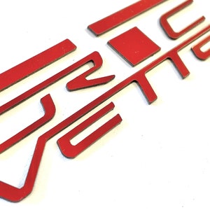 May include: Red metal letters spelling out the word "Vette" in a stylized font. The letters are cut out and have a reflective finish.