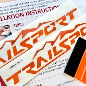 Könnte beinhalten: Orange-weiße "TRAILSPORT" Aufkleber mit Bergdesign, zusammen mit einem schwarzen und orangefarbenen Rakel. Installationsanweisungen sind im Hintergrund sichtbar.