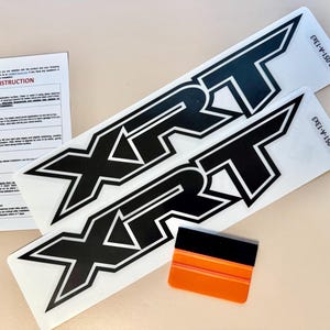 May include: Two black and white XRT decals with a small orange and black applicator tool and an installation instruction sheet. The decals are rectangular with the letters XRT in a bold, stylized font. The installation sheet has step-by-step instructions.