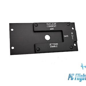 Może przedstawiać: Czarny metalowy panel z okrągłym przyciskiem i prostokątnym wycięciem. Panel zawiera tekst "NUCLEAR CONSENT ARM", "SAFE", "NUC" i "JETTISON ENABLE".