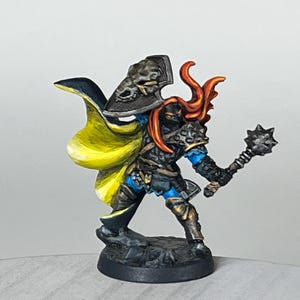 May include: A miniature figurine of a warrior in detailed armor, holding a spiked mace. The figure has a yellow and black cape, a shield with a skull design, and orange hair. The armor is painted in shades of black, blue, and gold.