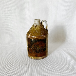 Puede incluir: Una jarra de cerámica marrón vintage con asa y una etiqueta descolorida que representa una escena de personas reunidas alrededor de una mesa. La jarra tiene un aspecto desgastado y rústico.