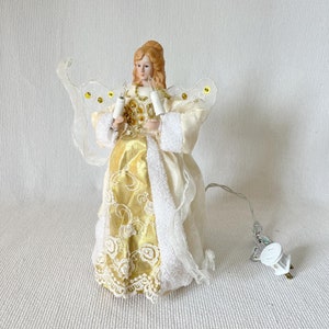May include: A Christmas tree topper in the shape of an angel. The angel has blonde hair and is wearing a cream-colored dress with gold accents and lace detailing. She holds two white candle lights and has sheer wings with gold sequins.