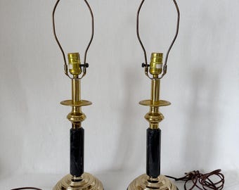 May include: Pair of vintage table lamps with brass bases and accents, black marble columns, and brown lamp cords. Each lamp has a brass harp and a bulb socket. The lamps are on a white background.