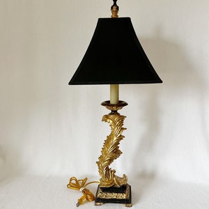May include: A gold table lamp with a black lampshade. The lamp base is decorated with a swirling leaf design. The lamp has a black square base.