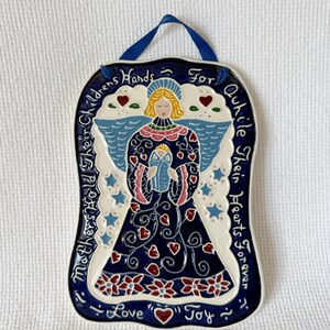 May include: A ceramic wall hanging featuring an angel holding a baby, with blue wings and a navy blue dress. The design includes red hearts, stars, and floral accents. The text reads "Mothers Hold Their Children's Hands For Awhile Their Hearts Forever Love Joy".