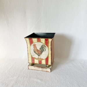 Puede incluir: Un macetero de metal blanco con rayas rojas y doradas. El macetero tiene un diseño de gallo en la parte delantera y un borde negro. El macetero se asienta sobre una pequeña base de madera.