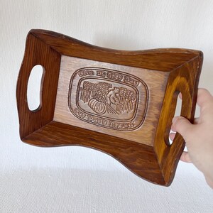 May include: A wooden serving tray with a carved design of wheat and a person harvesting. The tray has two handles and the text "Give us this day our daily bread".