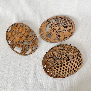 May include: Three oval-shaped copper trivets with intricate cut-out designs. One features a rose, another a basket of flowers, and the third a cluster of fruit.
