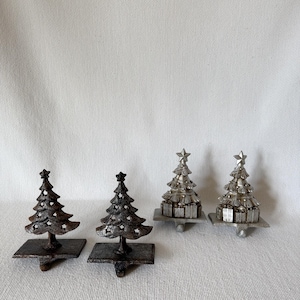 May include: A set of four metal stocking holders featuring Christmas tree designs. Two holders are dark brown with star cutouts, and two are silver-toned with a layered tree structure resting on a base of gift boxes, each topped with a star.