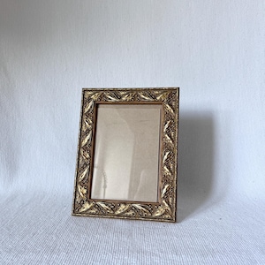 May include: An antique-style picture frame with a gold finish. The frame has a detailed, repeating leaf and floral pattern around the edges. The frame is empty, with a clear glass or plastic insert.