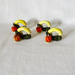 May include: Set of four ceramic napkin rings featuring a green squash and a small orange pumpkin. The rings are designed to hold napkins in place at a dinner table.